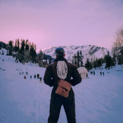 Back view anonymous male traveler in warm clothes standing with hands in pockets on snowy mountain slope in modern ski resort at twilight