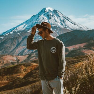 Young stylish man in hat and modern sunglasses standing on hill with hand in pocket against high snowy mountain